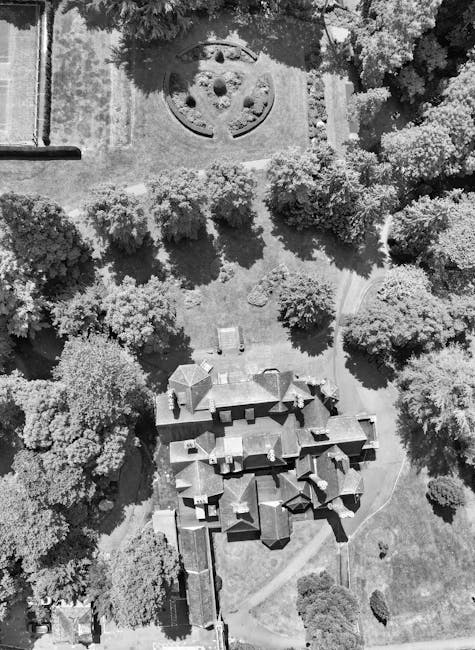 An aerial view of a garden scene showcasing a large, historic building with multiple pitched roofs and chimneys, surrounded by a well-maintained lawn and a variety of trees with dense foliage. A curve