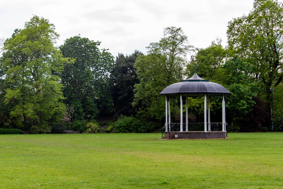 A wide view of a well-maintained park with a lush green lawn and a variety of mature trees, including both deciduous and evergreen species. In the center-right of the image, there is a small, open pav