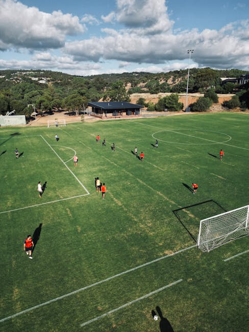 A wide view of a well-maintained park with a lush green lawn and a variety of mature trees, including both deciduous and evergreen species. In the center-right of the image, there is a small, open pav